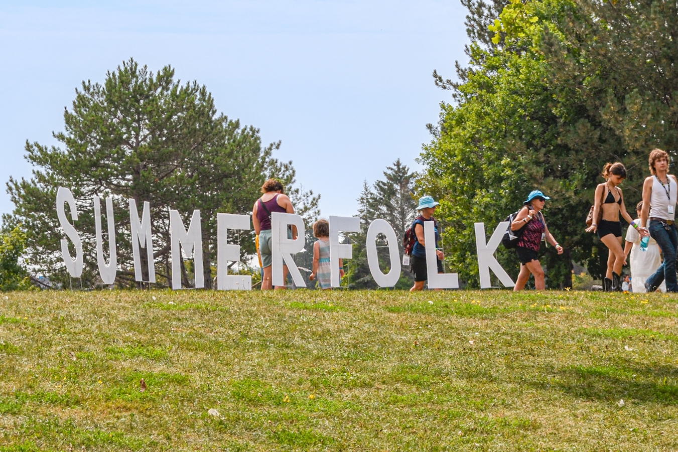 Festival goers walk over a grassy hill. Large white letters spelling "SUMMERFOLK" stand on the hill.