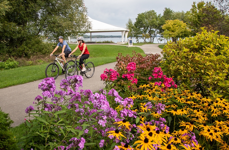Two cyclists riding along a paved waterfront path, passing bright flower beds with a white pavilion and trees in the background.