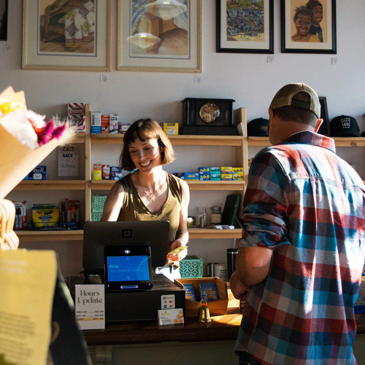 Customer stands at a small shop counter while a cashier processes a purchase, with shelves and framed artwork behind them.