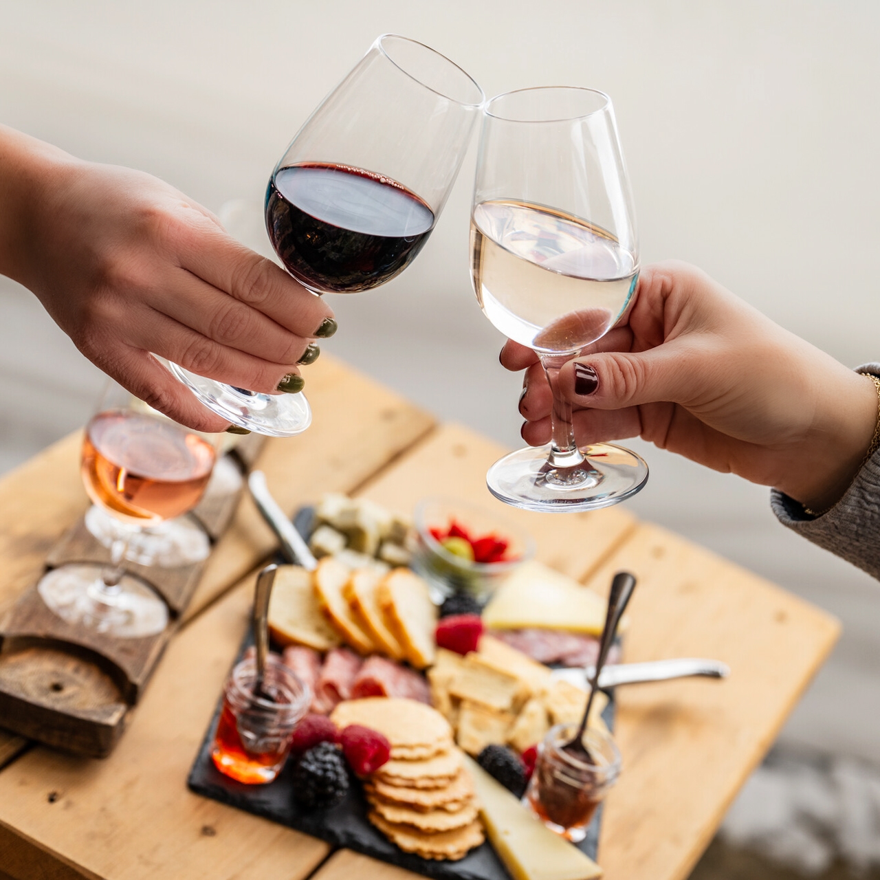 Two wine glasses raised over a wooden table with a charcuterie board of cheese, crackers, and fruit.