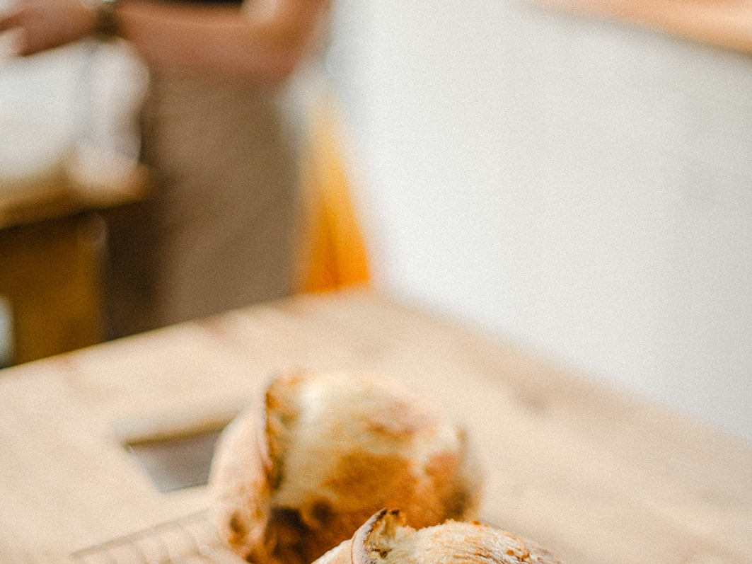Two loaves of sourdough in the foreground on a wooden table, with a woman in the background weighing ingredients out.