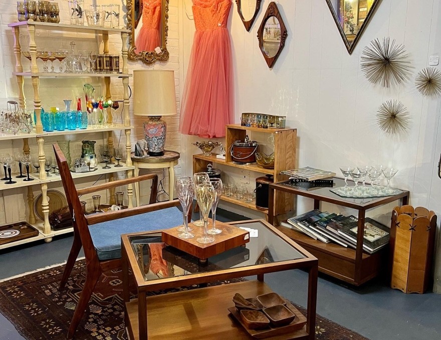 Picture of corner of shop with pink dress and mirrors hanging on wall with other decorative items, shelving with glasses, books and other items for sale with a chair and small glass table in the centre of the space.