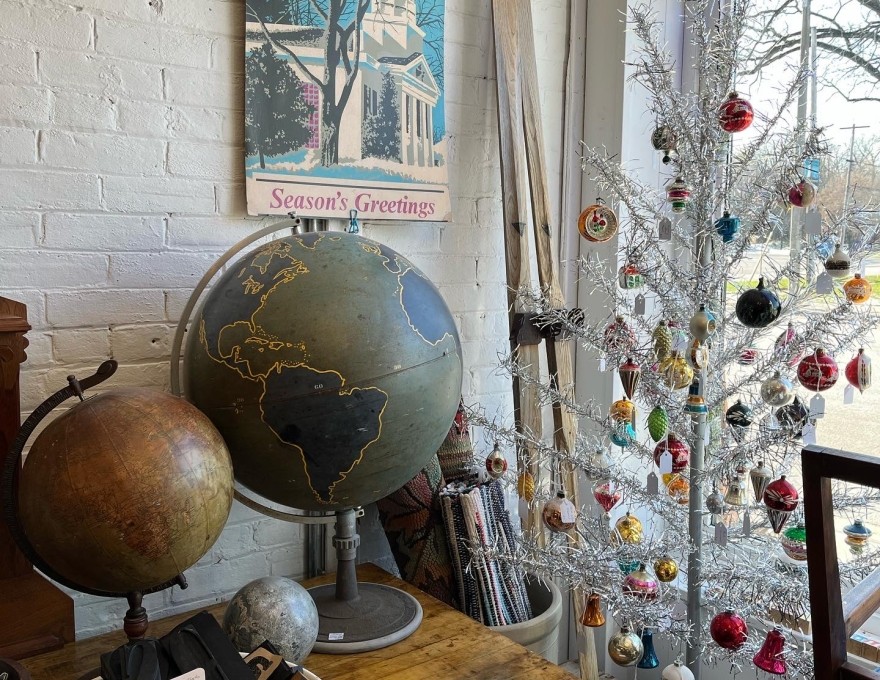 Picture of front window of shop from the interior looking outward with antique globes on tables, a &quot;Season's Greetings&quot; poster on the wall and a silver decorated Christmas tree in the window.