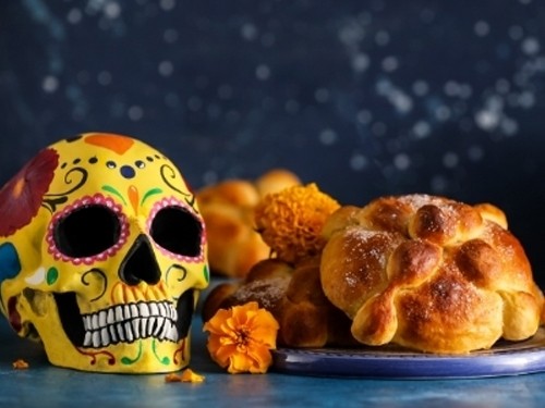 A colorful sugar skull decorated with vibrant patterns sits beside traditional Mexican "Pan de Muerto" pastries, surrounded by marigold flowers. The background is dark blue with white speckles resembling stars.