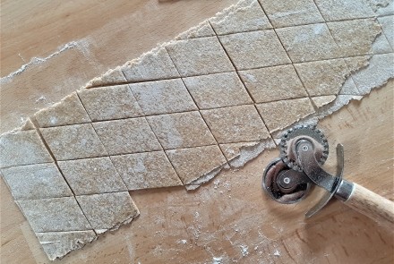 A sheet of pasta dough on a wooden counter, cut into diamonds, with a wooden handled pastry cutter laying on its side.