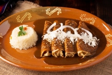 Four enchiladas topped with mole sauce, white cheese, and cream, served with white rice on a decorative brown ceramic plate over a woven placemat
