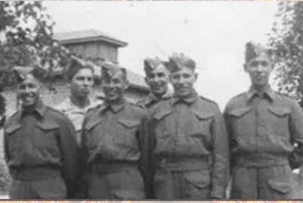 Historic black-and-white photo of five uniformed soldiers standing in a row outdoors, with a stone building and trees in the background.