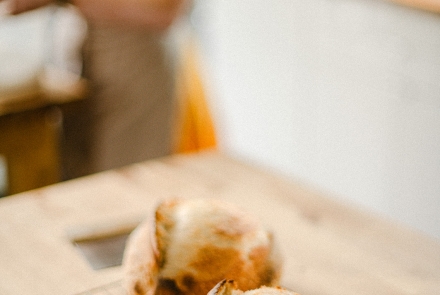 Two loaves of sourdough in the foreground on a wooden table, with a woman in the background weighing ingredients out.