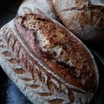 A rosy amber baked loaf of sourdough, with a score down the centre of the oblong shape, and leaf-like scoring down the left side.