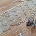 A sheet of pasta dough on a wooden counter, cut into diamonds, with a wooden handled pastry cutter laying on its side.