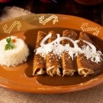 Four enchiladas topped with mole sauce, white cheese, and cream, served with white rice on a decorative brown ceramic plate over a woven placemat