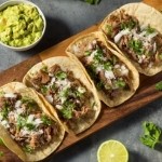 Four tacos filled with shredded meat, onions, and cilantro on a wooden board, served with guacamole, lime wedges, and scattered cilantro leaves.